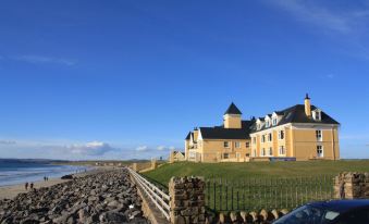a large , yellow house with a black roof and black chimney is situated on a grassy beach , under a clear blue sky at Sandhouse Hotel