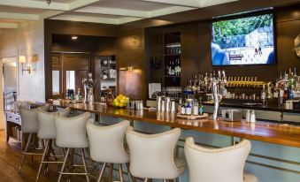 a bar area with several white chairs and a television mounted on the wall , surrounded by bottles and cups at Shady Villa Hotel