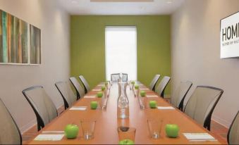 a long wooden table with green apples and glass cups is set up in a room with chairs at Home2 Suites by Hilton Dekalb