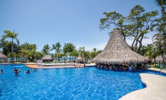 a large blue pool surrounded by a resort with palm trees and a thatched - roof hut at Barcelo Tambor - All Inclusive