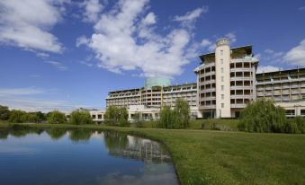 a large white building surrounded by a grassy field and a body of water , possibly a lake or a pond at UNA Hotels Expo Fiera Milano