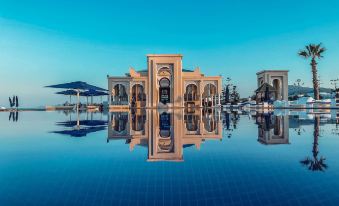 a large pool with a traditional building in the background , reflecting in the water and surrounded by umbrellas at Banyan Tree Tamouda Bay