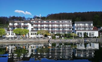 a large white building with a reflection in the water , surrounded by trees and mountains at Seehotel Leoni