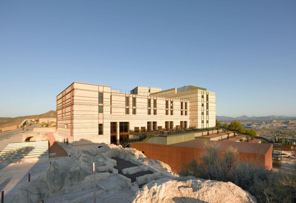 a modern building with a stone wall and large windows is situated on a rocky outcropping at Parador de Lorca