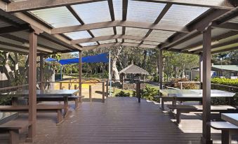 a covered outdoor area with wooden flooring , picnic tables , and umbrellas , surrounded by trees and buildings at Discovery Parks - Emerald Beach