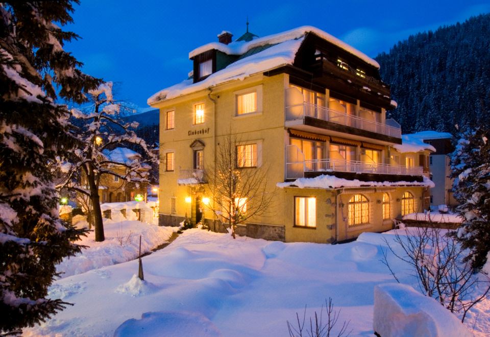 a large , yellow house with a balcony and lit windows is surrounded by snow - covered trees at Hotel Lindenhof