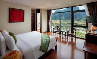 a large bed with a white comforter and green throw is in a room with wooden floors , chairs , and a window overlooking a scenic landscape at Tam Coc La Montagne Resort & Spa