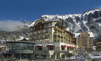 a large , multi - story hotel surrounded by snow - covered mountains , with a restaurant on the ground floor at Arenas Resort Victoria-Lauberhorn