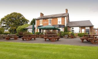 a brick building with an open - air restaurant area featuring wooden benches and tables , surrounded by greenery at Premier Inn Crewe (Nantwich)