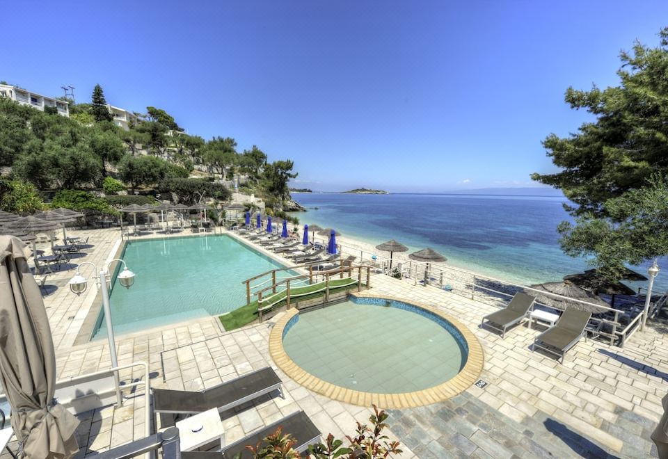 a large outdoor swimming pool surrounded by lounge chairs and umbrellas , with a view of the ocean in the background at Agali Hotel Paxos