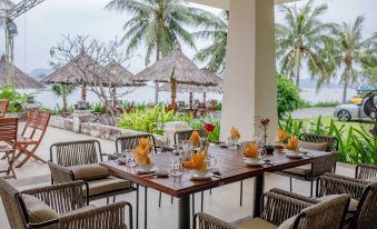 a restaurant patio with wooden tables , chairs , and umbrellas , surrounded by palm trees and overlooking the ocean at Que Toi Village Resort Phu Yen