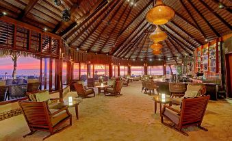 a large , open room with a thatched roof is furnished with wooden tables , chairs , and other furniture at Komandoo Island Resort & Spa