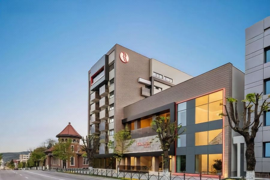 a modern hotel building with a red and white sign on the front , located in a city setting at Ramada by Wyndham Ramnicu Valcea