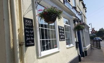 a building with a window display of food and drinks , a sign for food dairy , and two hanging flower baskets at The Lamb Inn