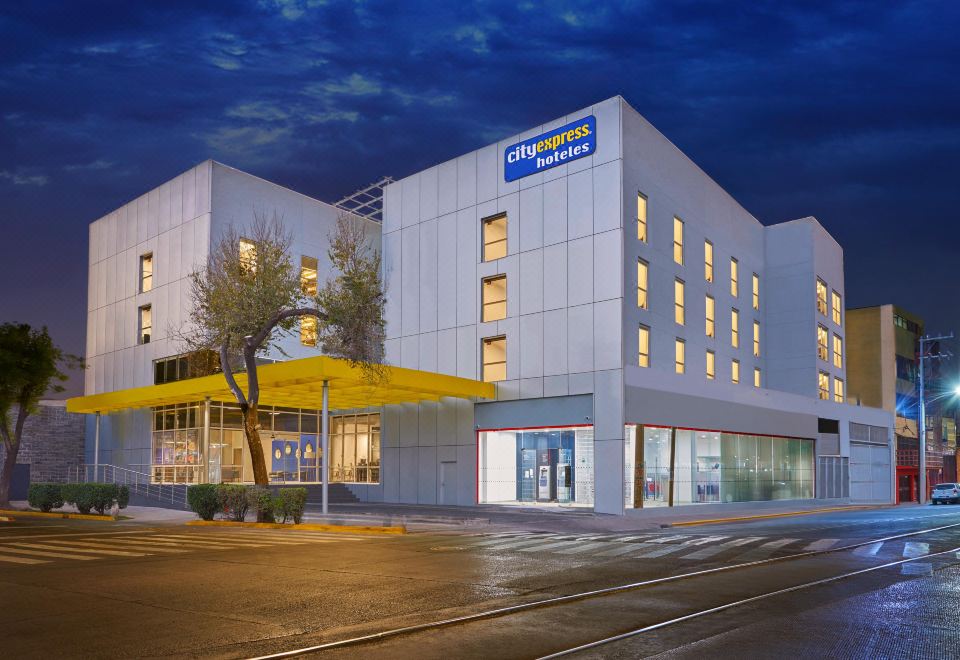 a modern hotel building with a yellow awning , located on a street corner at dusk at City Express by Marriott Cdmx la Villa