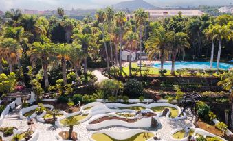 a lush tropical garden with palm trees , flowers , and a swimming pool surrounded by lush greenery at Precise Resort Tenerife