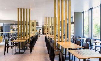 a modern , minimalist restaurant with wooden tables and chairs , large windows , and gold - colored pillars at Ibis Baden Neuenhof