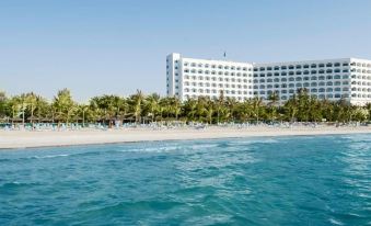 a large white building on the beach , with palm trees in the foreground and a clear blue sky above at Ajman Hotel by Blazon Hotels