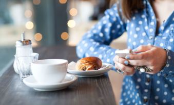 a woman in a blue polka dot dress is holding a croissant while sitting at a dining table with a cup of coffee at Sonesta Select Raleigh Durham Airport Morrisville