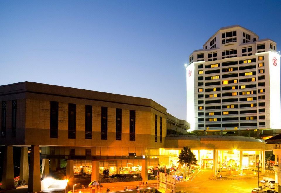 a tall building is lit up at night , with a parking lot in front of it at Thumrin Thana Hotel