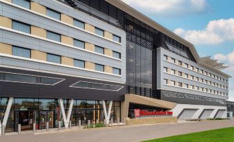 a modern building with large windows and a gray roof , surrounded by green grass and trees at Hilton Garden Inn Silverstone