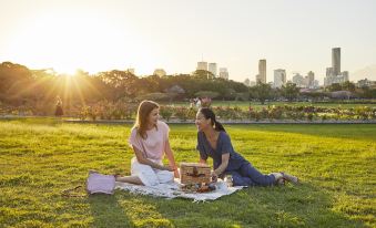two women are sitting on a blanket in a grassy field , enjoying a picnic with a view of the city in the background at Quest Cannon Hill
