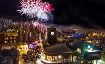 a nighttime scene of a snowy village with a large group of people gathered around a clock tower , watching fireworks in the sky at Sundance Resort