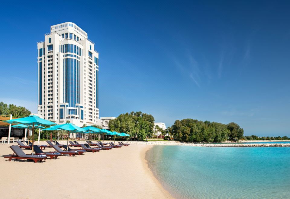 a tall white building with blue windows is overlooking a sandy beach with lounge chairs and umbrellas at The Ritz-Carlton, Doha
