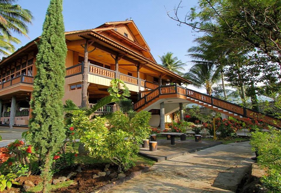 a large wooden house with a porch and balcony is surrounded by greenery and palm trees at Danau Dariza Resort Hotel - Cipanas Garut