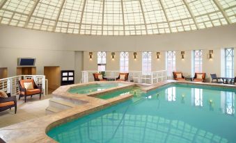 an indoor swimming pool with a glass ceiling , surrounded by lounge chairs and tables , in a well - lit room at Omni Providence Hotel