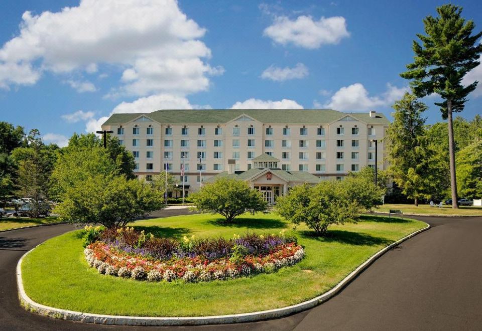 a large hotel building surrounded by a lush green lawn , with trees and flowers in the foreground at Hilton Garden Inn Albany