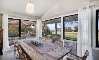 a wooden dining table surrounded by chairs in a room with large windows overlooking a field at Bear Gully Coastal Cottages