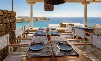 a dining table set with plates and utensils , surrounded by chairs on a wooden deck overlooking the ocean at The Summit of Mykonos