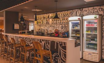 a modern bar with a black and white patterned wall , wooden chairs , and a refrigerator filled with various bottles at Ibis Carlos Barbosa