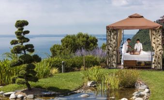 a man and a woman are sitting on a bed , surrounded by lush greenery , overlooking a body of water at Lefay Resort & Spa Lago di Garda