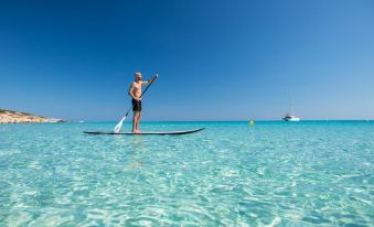 a man is standing on a paddle board in the ocean , holding a paddle and enjoying his time at Insotel Hotel Formentera Playa