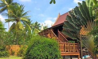 a wooden house surrounded by lush green grass and palm trees , with a swimming pool in the background at BaanSuanLeelawadee Resort Amphawa