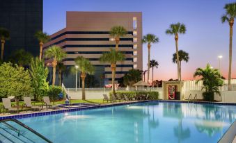 an outdoor pool surrounded by palm trees , with a building in the background and people enjoying their time near the pool at Hilton Melbourne, FL