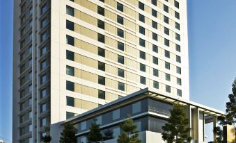 a tall white building with a green roof , located in a city setting with trees and clear skies at Pullman at Sydney Olympic Park