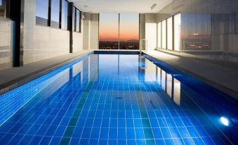 a long , empty swimming pool with blue tiles and large windows offering views of the city at sunset at Mantra Melbourne Airport