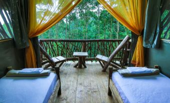 a wooden deck with two chairs , a table , and yellow curtains is shown in front of a forest at La Tigra Rainforest Lodge