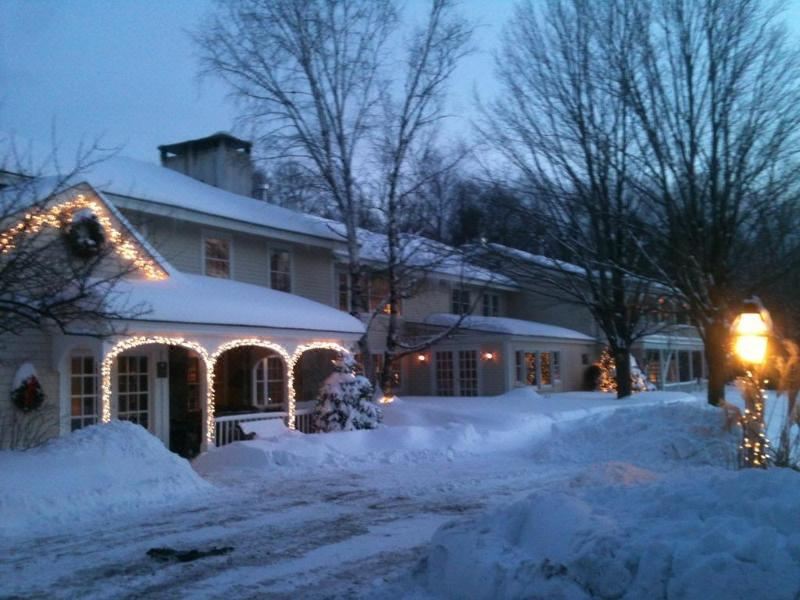 a snow - covered street with a house and trees , decorated with christmas lights and surrounded by snow at Deerhill Inn