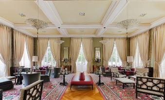 a spacious , elegant living room with high ceilings , large windows , and multiple seating arrangements , decorated with red cushions on the floor and white curtains at Vidago Palace