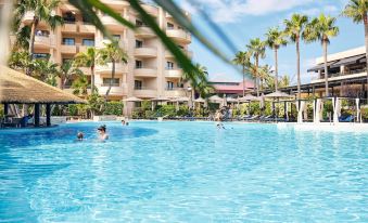 a large swimming pool with people in it and palm trees surrounding the building , under a clear blue sky at Protur Biomar Sensatori Resort