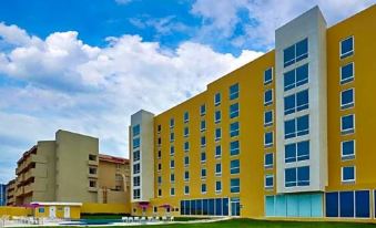 a large yellow and white building with blue windows , surrounded by green grass and a blue swimming pool at City Express by Marriott Nogales