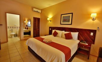 a large bed with a red and white comforter is in a hotel room with a bathroom visible through a doorway at Maun Lodge