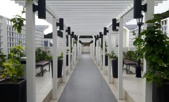 a long , white walkway with black poles and black planters is lined with benches under a covered area at G Residence