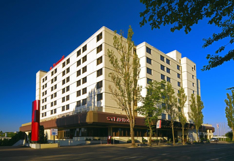 a tall building with a red sign on the front is surrounded by trees and a sidewalk at Stuttgart Marriott Hotel Sindelfingen