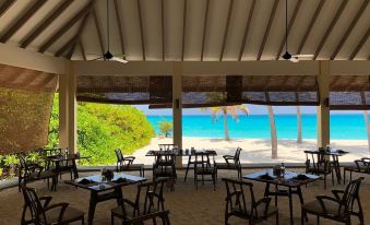 a restaurant with a large dining table surrounded by chairs , located near the beach and overlooking the ocean at Hondaafushi Island Resort