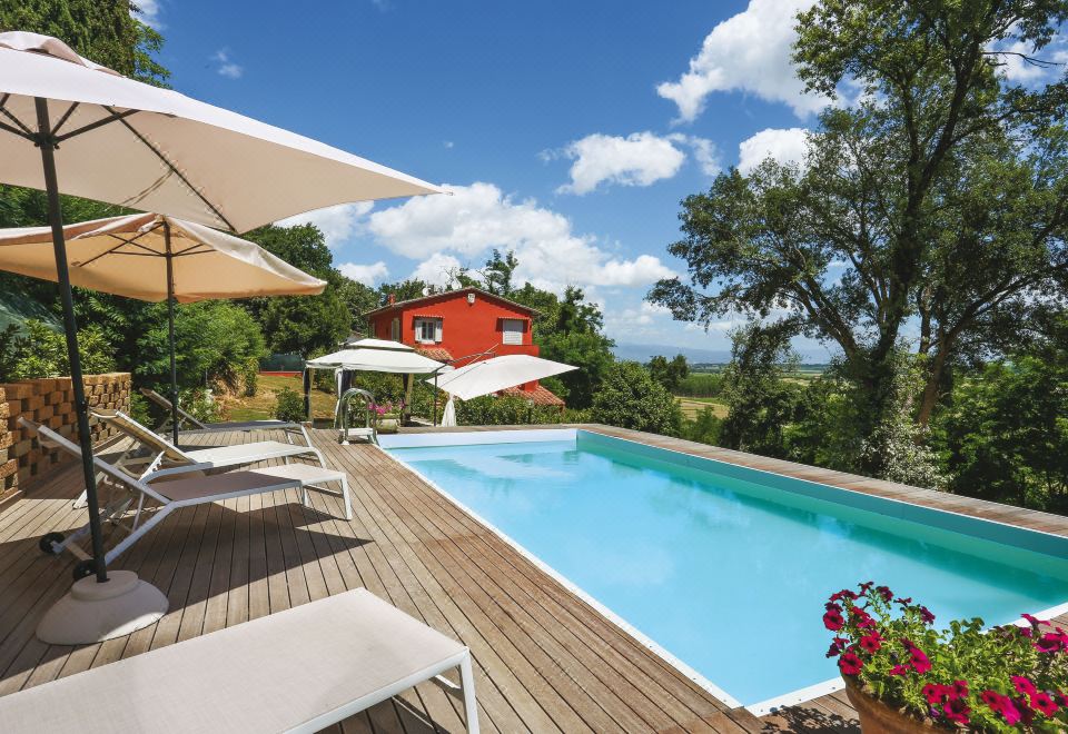 a large swimming pool is surrounded by lounge chairs and umbrellas , with a red house in the background at Piero